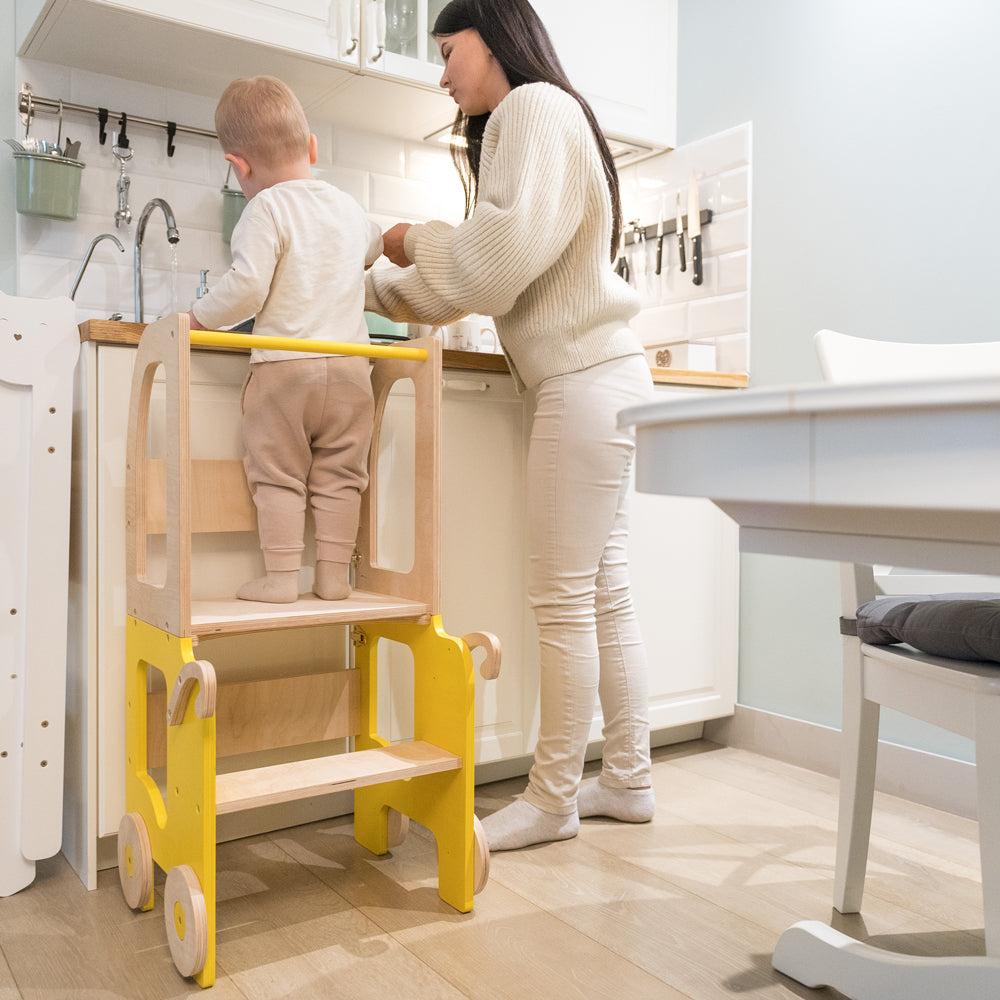 Kind steht auf 2in1 Lernturm im Auto-Look, unterstützt Spiel & Bewegung, ideal für gelbes Kinderzimmer und Montessori Spielmöbel Sammlung.