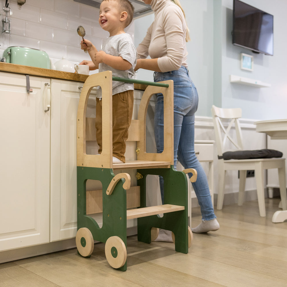 Kind steht auf umbaubarem 2in1 Lernturm im Auto-Look, unterstützt von Erwachsener in moderner Küche, Sitzmöbel für Kinder, Spiel & Bewegung Kinder.