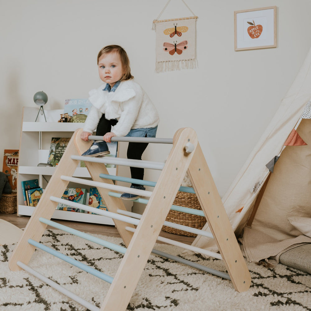 Kind klettert auf hellem Holz Kletterdreieck in pastellfarbenem Kinderzimmer mit Teppich und Spielbüchern im Hintergrund