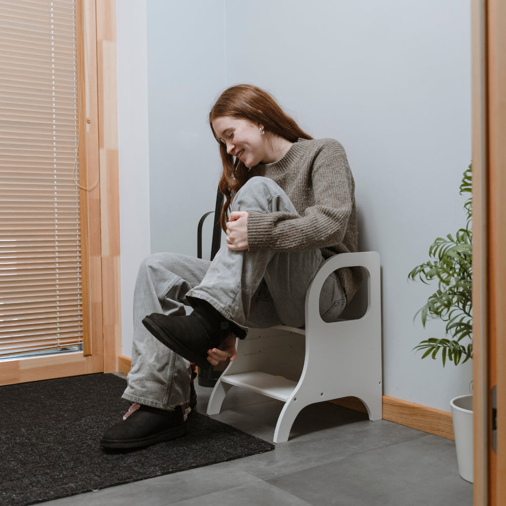 Frau sitzt entspannt auf höhenverstellbarem weißem Tritthocker aus Holz, ideal für Selbstständigkeit und stabile Unterstützung im Alltag.