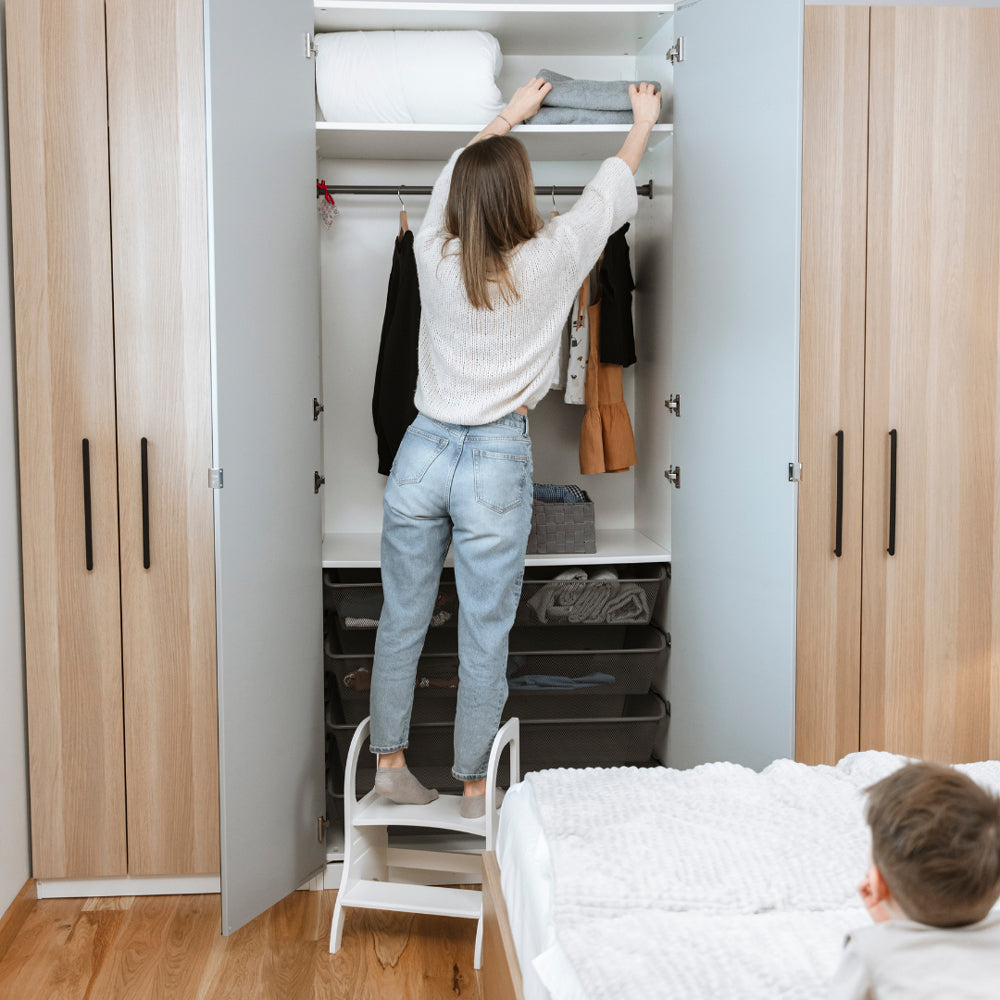 Frau nutzt höhenverstellbaren weißen Tritthocker aus Holz, um Kleidung im hohen Schrank zu erreichen, nachhaltiges Kinder-Tritthocker-Design.