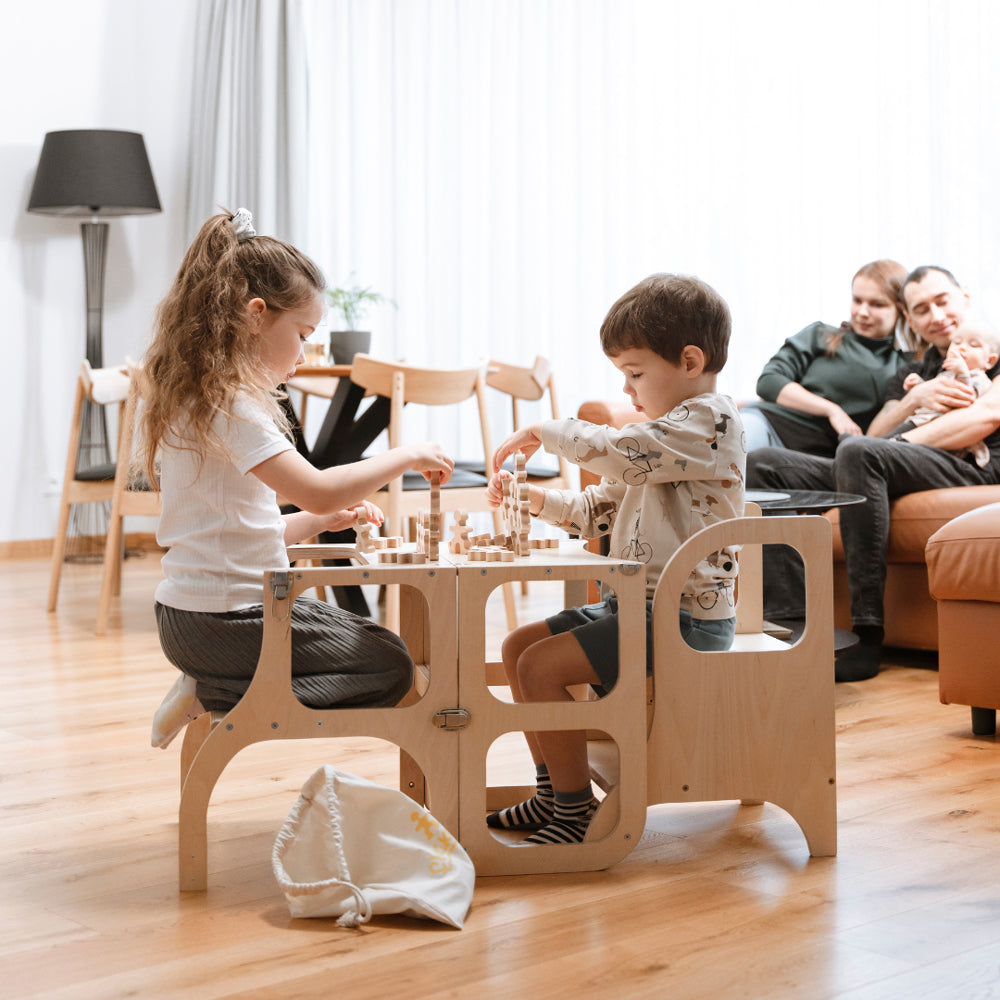 Kinder spielen an einem stabilen Holz-Tritthocker mit verstellbaren Stufen, während Eltern im Hintergrund entspannt zuschauen.