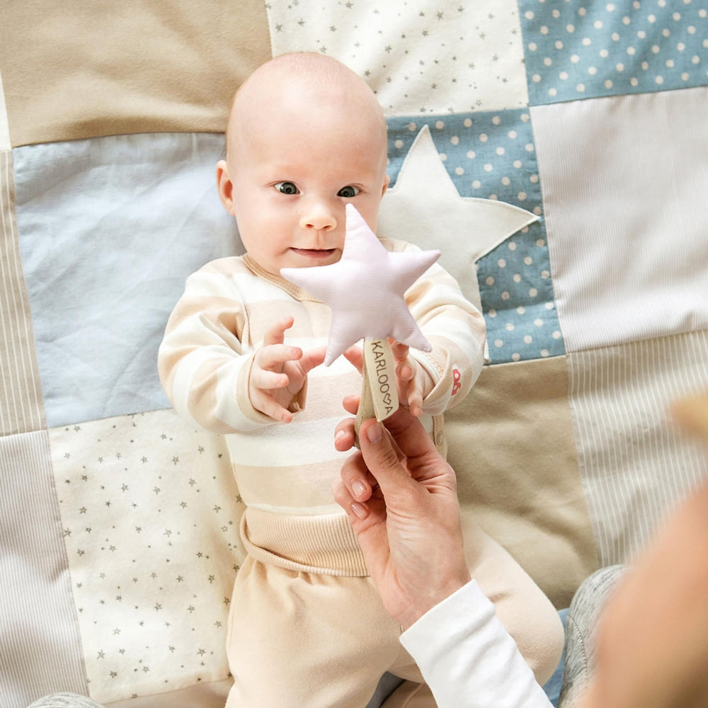 Baby liegt auf einer blau-beigen Patchwork Spieldecke und greift nach einem weichen Stern mit Rassel am Spielbogen.
