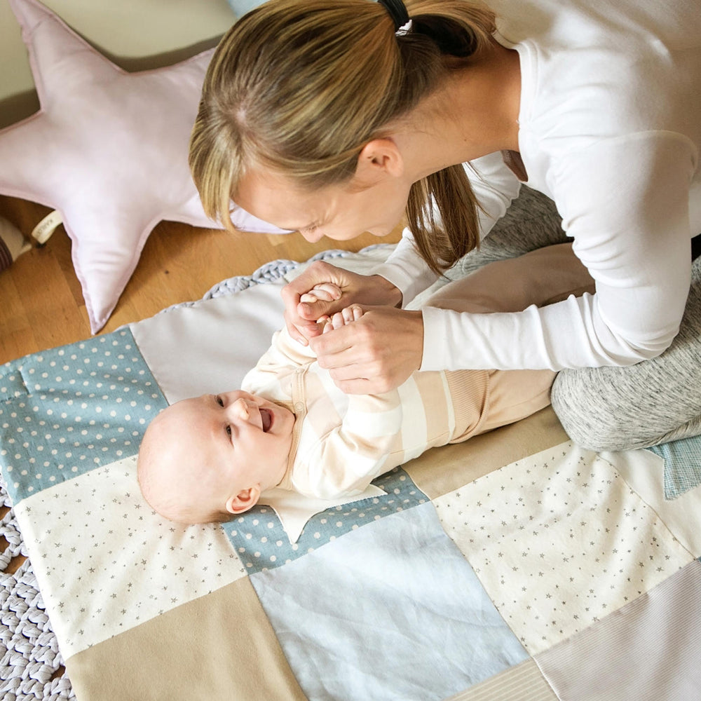 Baby liegt lachend auf einer blauen Patchwork-Spieldecke mit Spielbogen, sanfte Farben und spielerische Förderung der Motorik.