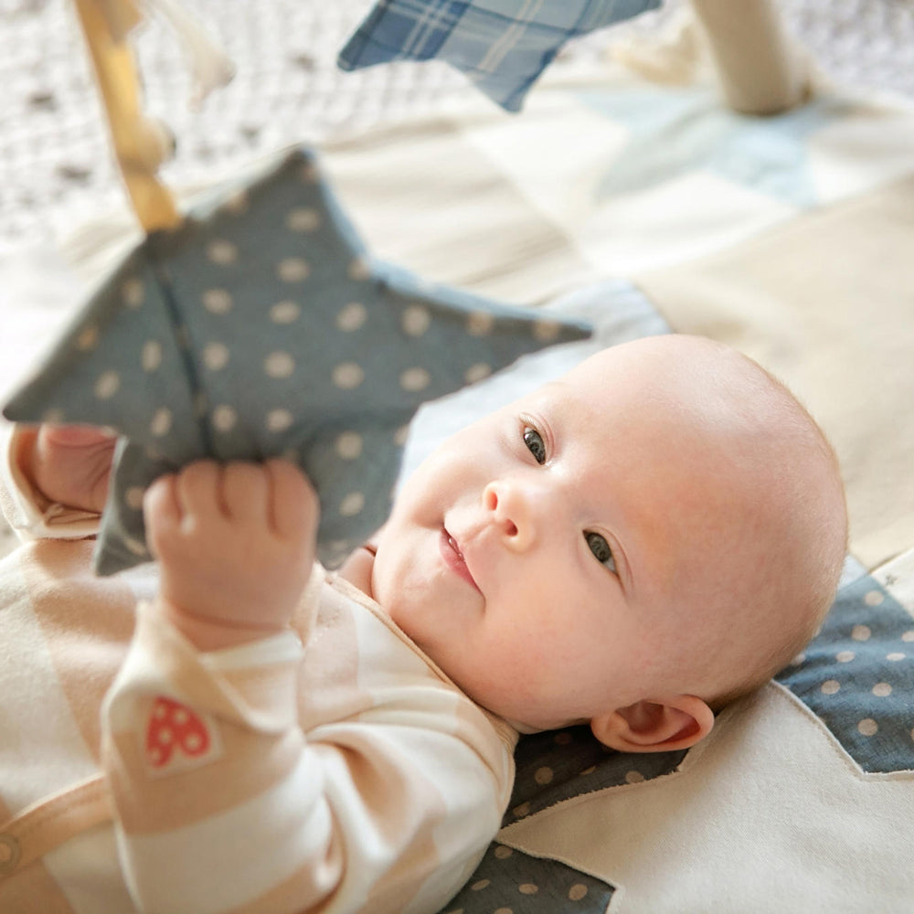 Baby liegt auf blauer Patchwork-Spieldecke und erkundet spielerisch weiche, hängende Sterne am abnehmbaren Spielbogen.