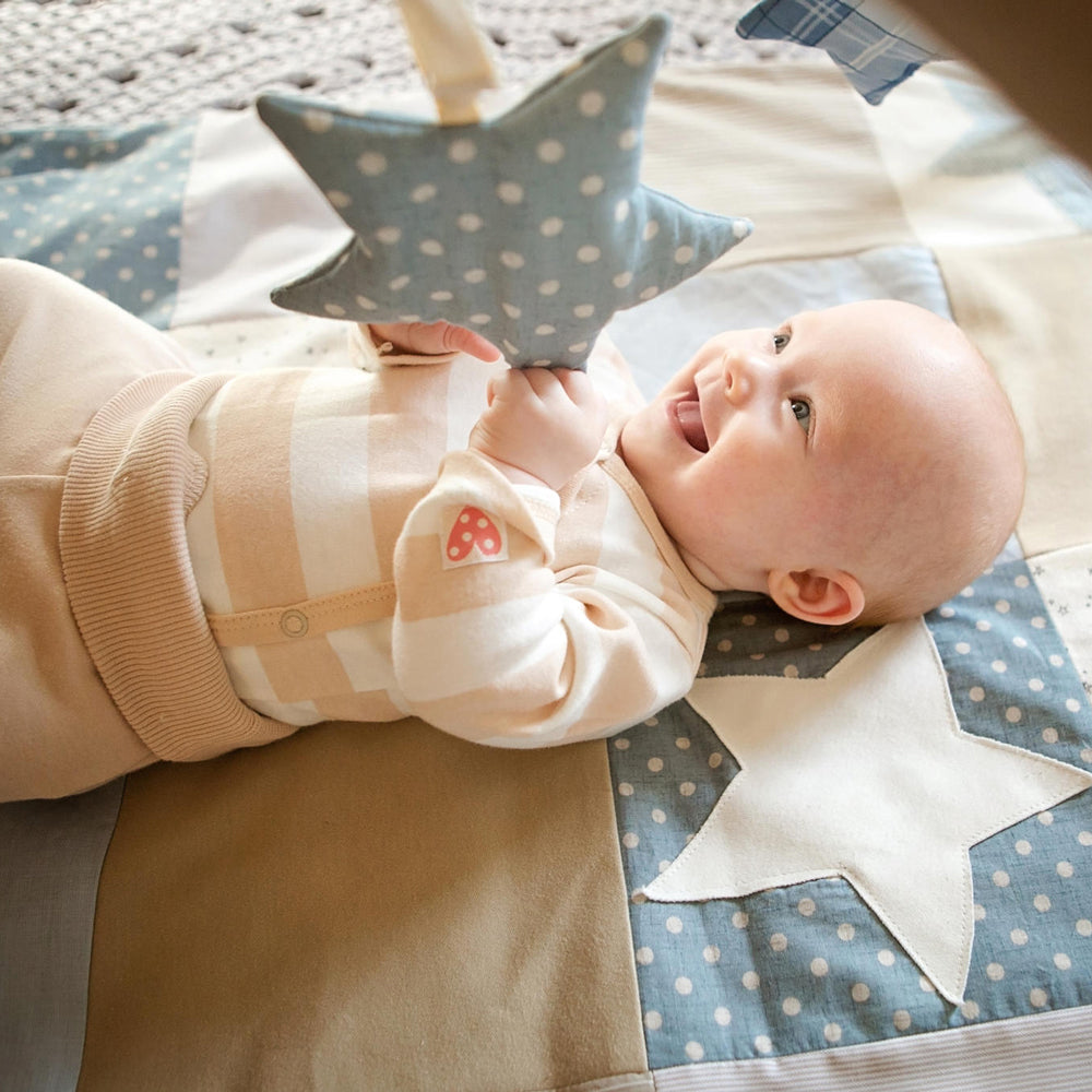 Glückliches Baby liegt auf patchwork Krabbeldecke in Blau und greift nach blauem Stern mit Punkten am Spielbogen, spielerisch Motorik fördern.