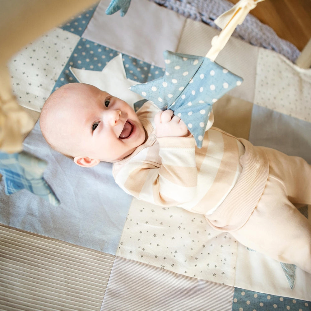 Glückliches Baby spielt auf weicher Activity Spieldecke Patchwork mit blauem Spielbogen und sterneförmigen Spielzeugen zur Förderung der Motorik.