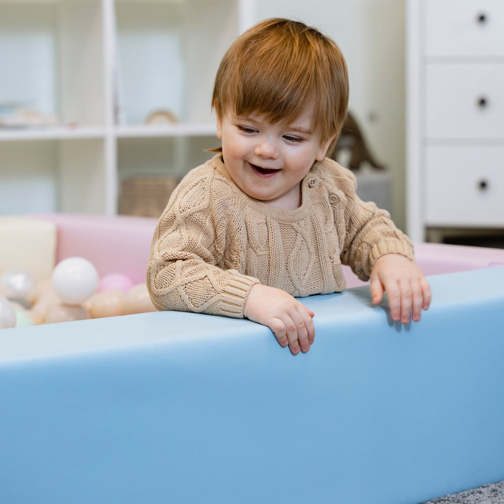 Glückliches Kleinkind spielt im quadratischen Bällebad aus hellblauem, weichem Kunstleder, sicherer Spielplatz für Entdecker ab 9 Monaten.
