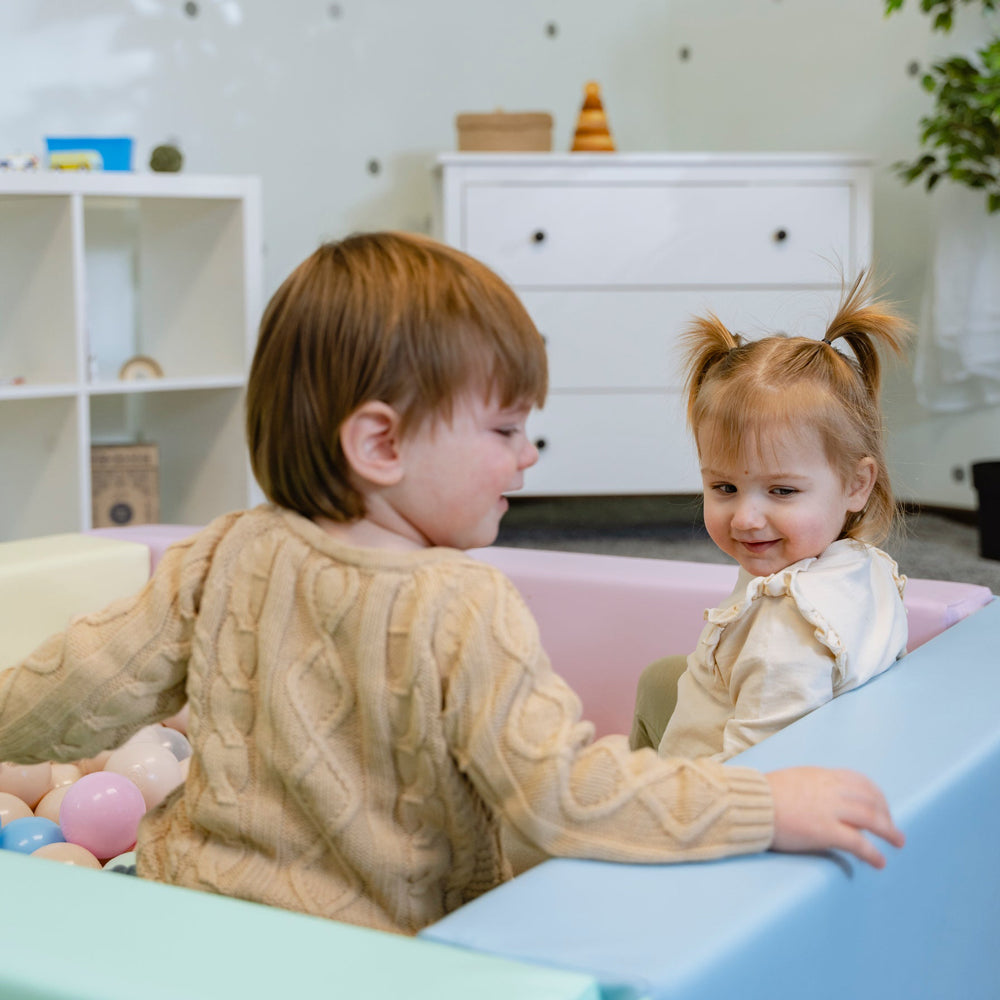 Zwei Kleinkinder sitzen in einem pastellfarbenen quadratischen Bällebad aus Kunstleder und spielen fröhlich miteinander.