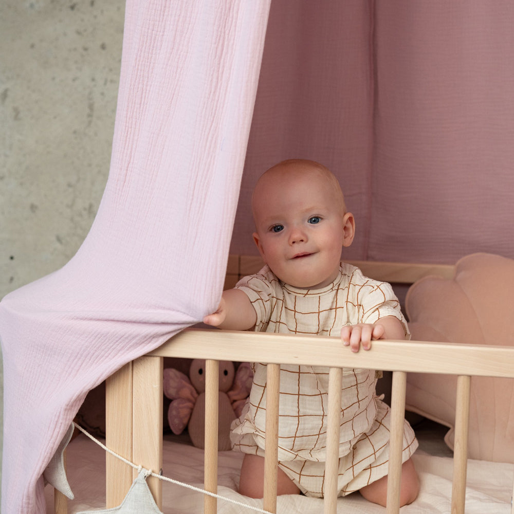 Baby sitzt im Kinderbett mit rosafarbenem Betthimmel aus Bio Baumwolle, gemütlicher Rückzugsort zum Träumen und Spielen.