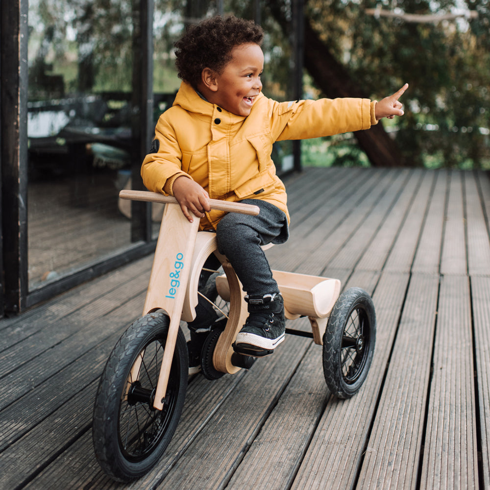 Kind auf dreirädrigem, modularem Laufrad mit Erweiterungsset, spielt fröhlich und sicher auf Holzterrasse im Freien.