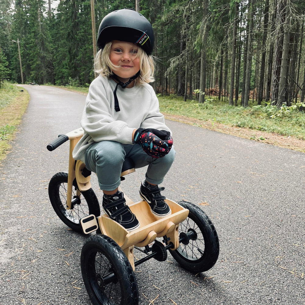 Kind sitzt sicher auf einem umgebauten 3in1 Laufrad mit Dreirad-Erweiterungsset auf Waldweg, trägt Helm und Handschuhe.