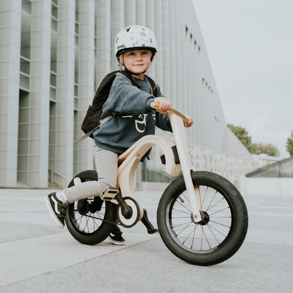 Kind mit Helm fährt umweltfreundliches 3in1 Laufrad mit Pedal-Erweiterungsset aus lackiertem Birkensperrholz und Metallrädern.