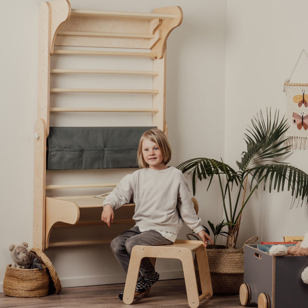 Kind sitzt auf kinderfreundlichem Holzstuhl neben angebrachtem Tisch an 2in1 Kletterdreieck mit natürlicher Maserung im Kinderzimmer.