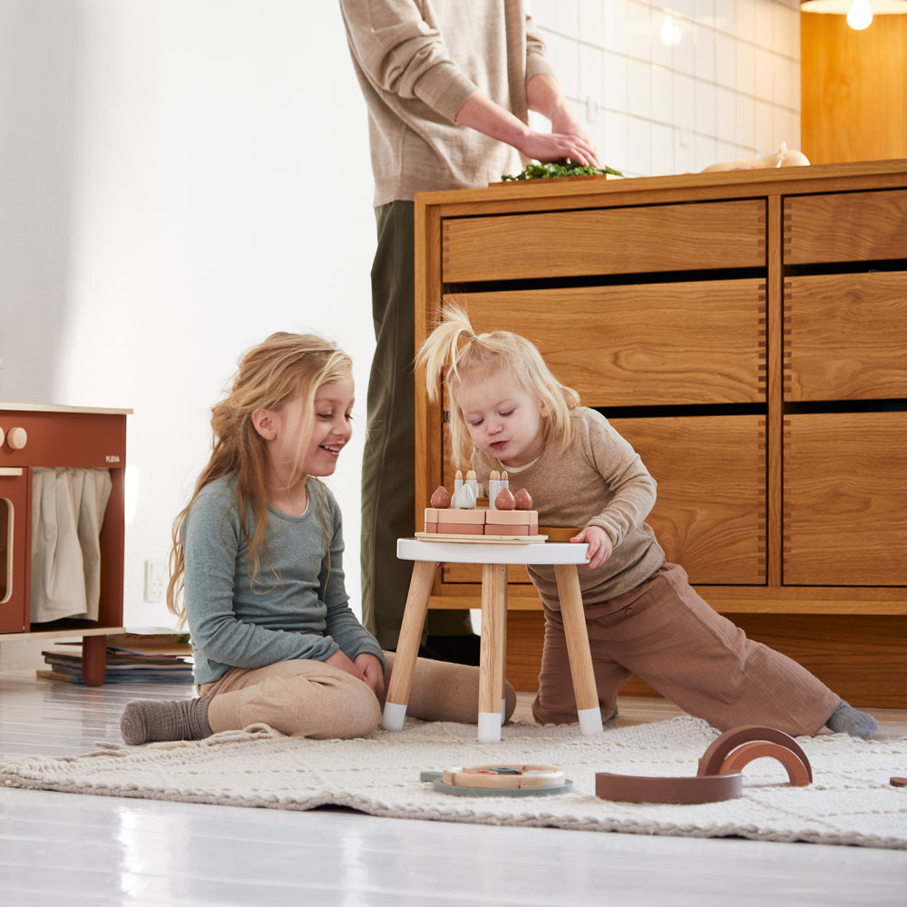 Kinder spielen mit dem Geburtstagskuchen-Spielset aus Holz für Kinderküchen, 25 Teile, auf einem Teppich im Wohnzimmer.