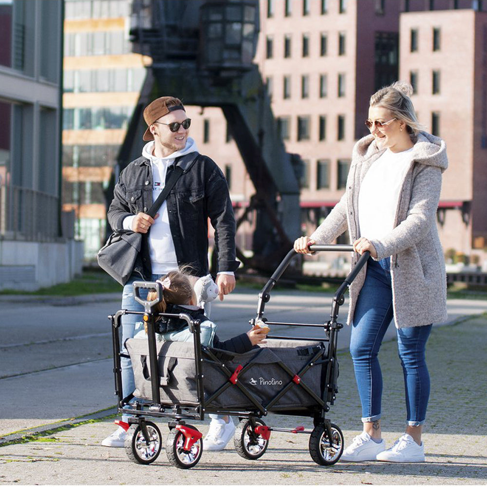 Junge Familie nutzt klappbaren Bollerwagen mit Sonnendach und Bremse aus der Kollektion Spielzeug & Spiele für Kinder draußen im urbanen Umfeld.