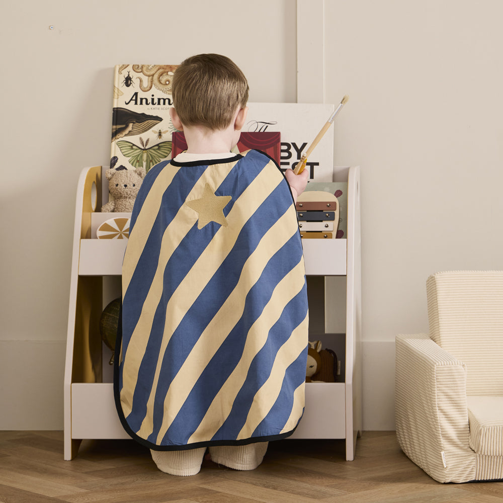 Kind mit blau-gelb gestreiftem Umhang mit Stern, spielt vor Regal mit Büchern und Spielzeug in Kinderzimmer mit gemütlichem Sessel
