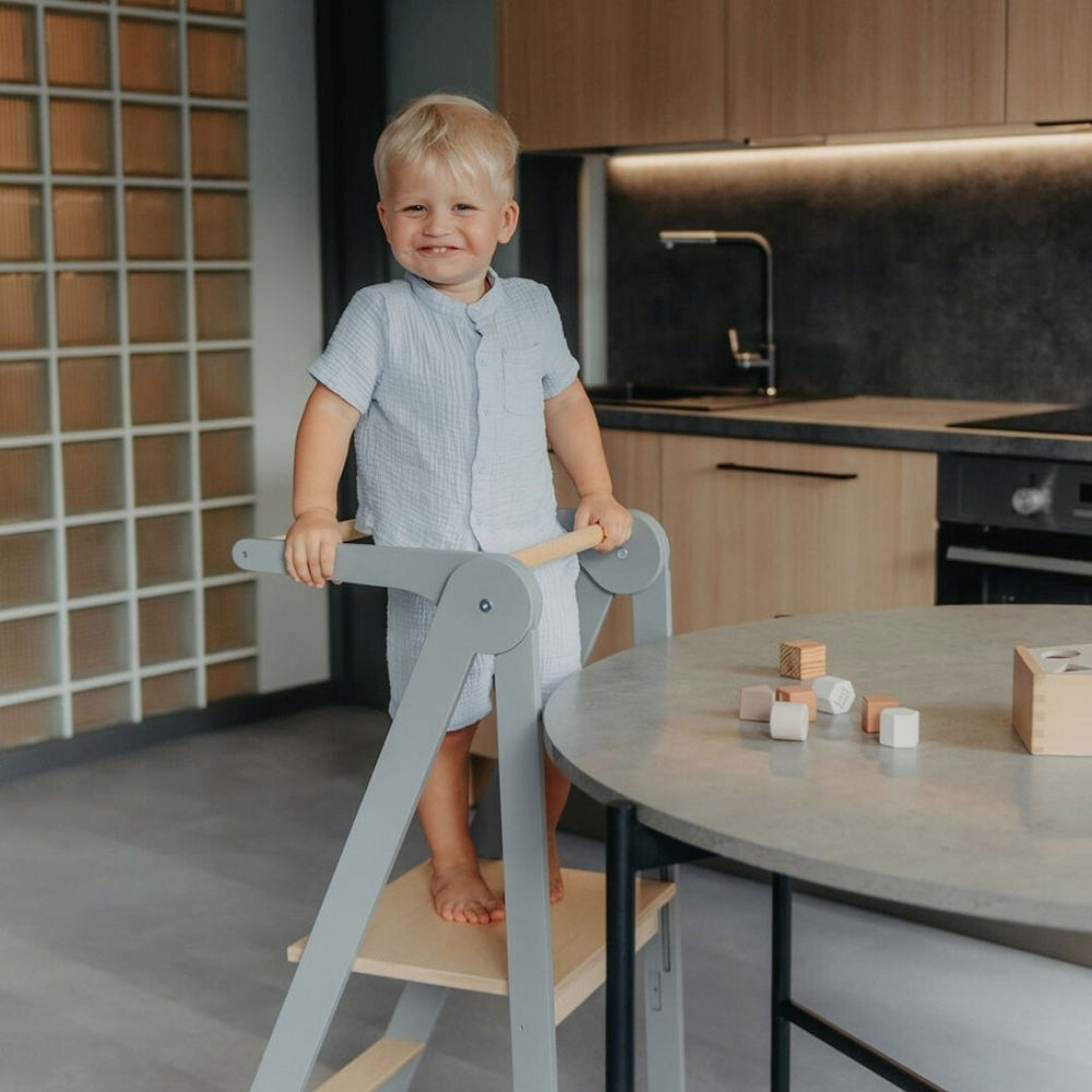 Kleiner Junge steht glücklich auf höhenverstellbarem und klappbarem Lernturm aus Holz aus der Sammlung Spielzeug & Spiele, Spielmöbel, Montessori.