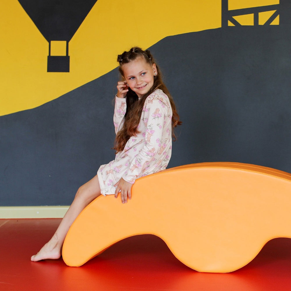 Mädchen spielt auf dem orangefarbenen Softblock Wippe Rocker aus der Kollektion Spielzeug & Spiele für Kinder Bewegung und Motorik.