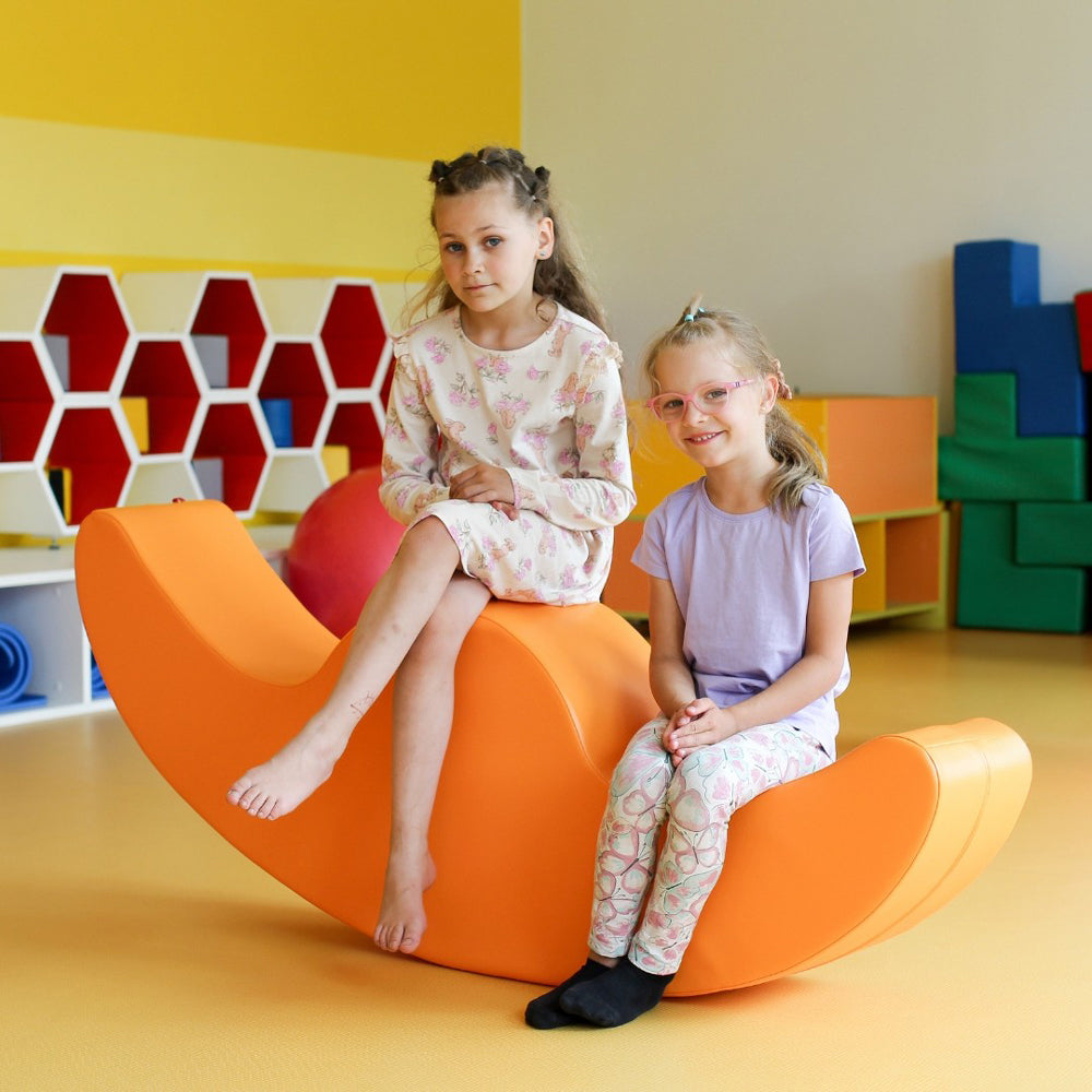 Zwei Kinder sitzen auf einem orangefarbenen Softblock Wippe Motorikspielzeug in einem hellen, freundlichen Spielzimmer für Kinder.