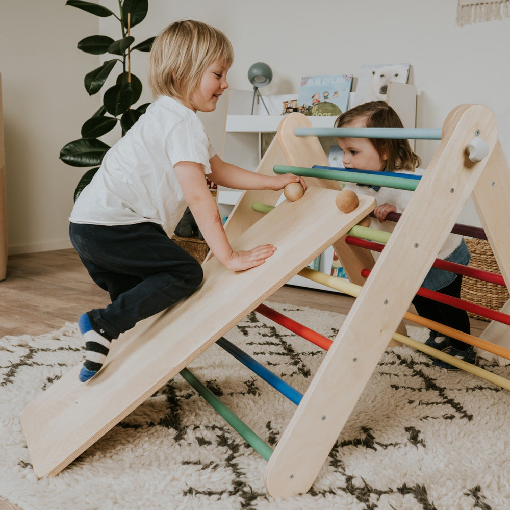 Zwei Kinder spielen mit einer Holzrampe und einem bunten Kletterdreieck im Kinderzimmer für Bewegung und Spaß.
