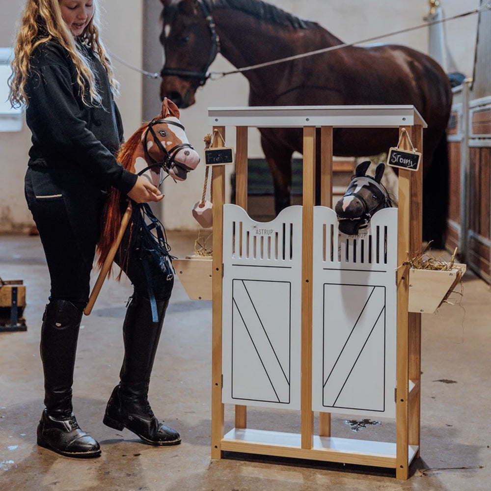 Kind spielt mit zwei Steckenpferden im liebevoll gestalteten Sperrholzstall aus der Sammlung Spielzeug & Spiele, Hobby Horsing.
