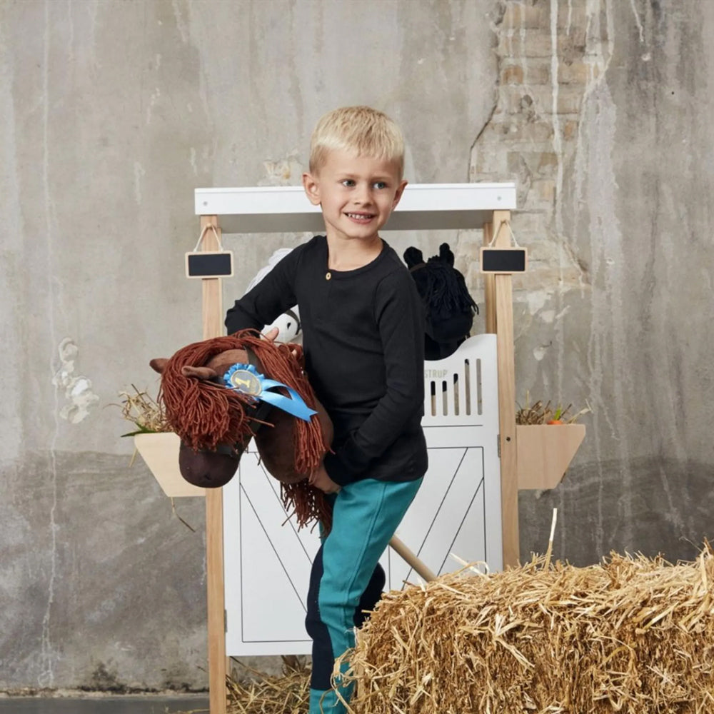 Kind mit braunem Steckenpferd aus der Hobby Horsing Tierwelt Themenwelt spielt fröhlich neben Heuballen in rustikalem Stall.