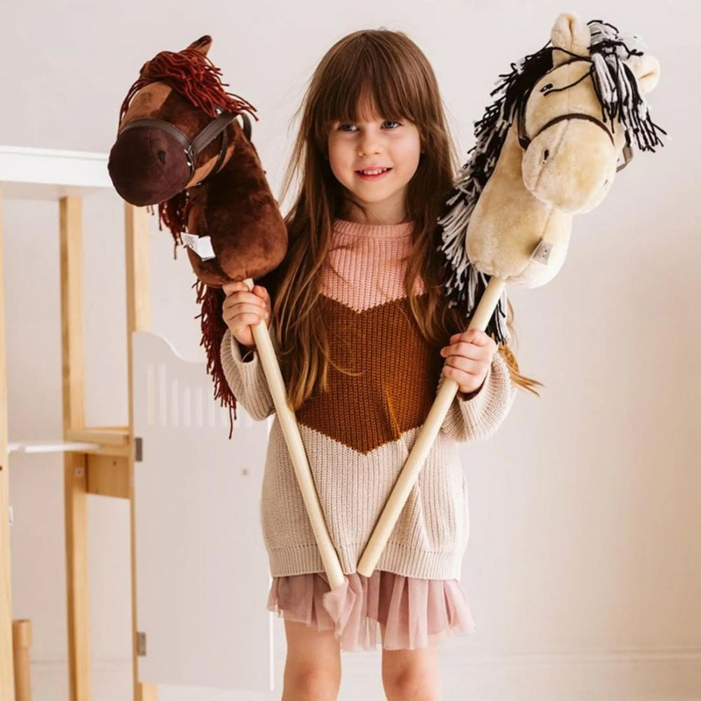 Mädchen hält zwei Steckenpferde in braun und beige aus der Hobby Horsing Tierwelt Themenwelt, fröhlich und verspielt.